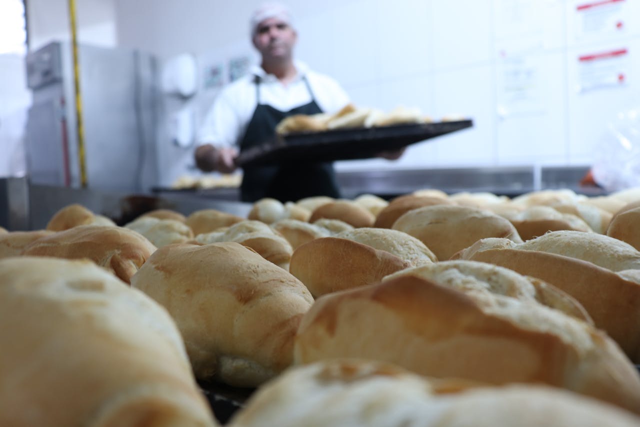 services-02 Close-up of freshly baked bread in a bakery kitchen, showcasing golden crusts and a baker in the background.